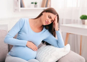 Mujer embarazada con dolor de cabeza y malestar estomacal, sentada en un sofá con expresión de incomodidad.