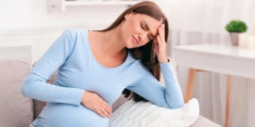 Mujer embarazada con dolor de cabeza y malestar estomacal, sentada en un sofá con expresión de incomodidad.