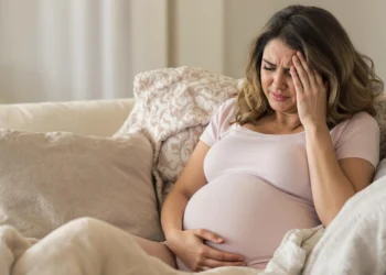 Mujer embarazada con dolor de cabeza y malestar, tocándose la frente mientras descansa en un sofá.