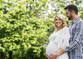 Pareja sonriente en un parque abrazando la barriga de embarazo de la mujer.