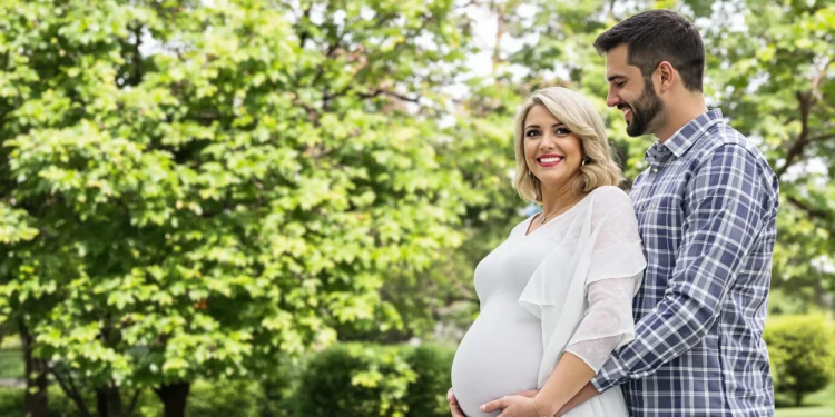 Pareja sonriente en un parque abrazando la barriga de embarazo de la mujer.
