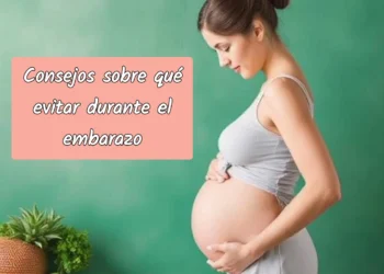 Mujer embarazada con vestido gris claro acariciando su vientre frente a una pared verde.