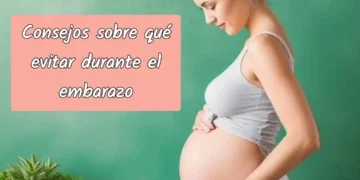 Mujer embarazada con vestido gris claro acariciando su vientre frente a una pared verde.